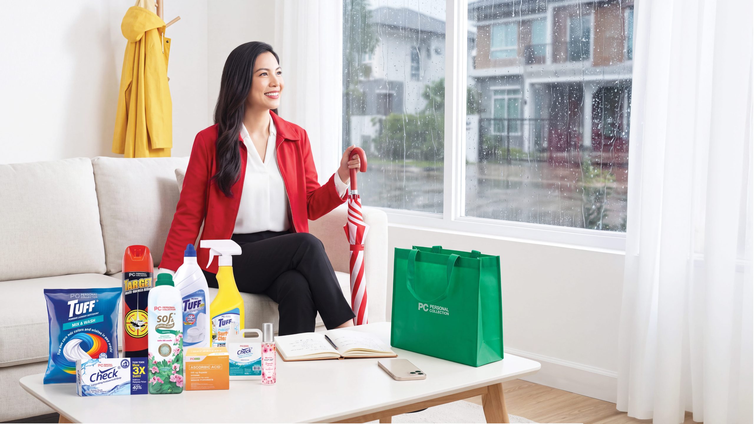 A woman in a red blazer sits indoors by a rainy window next to a collection of household cleaning supplies and a green shopping bag from Personal Collection.