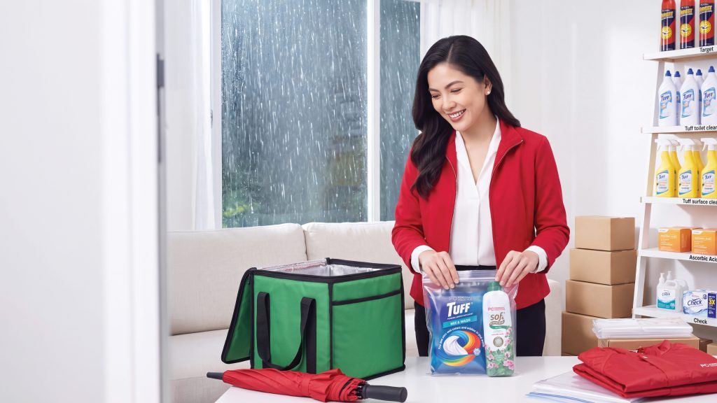 A smiling woman in a red jacket sealing a ziplock bag of Personal Collection laundry products next to a green insulated bag.