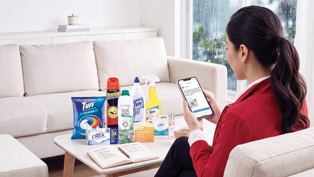 A woman sitting on a couch, looking at her phone next to a coffee table filled with Personal Collection household cleaning products and a notebook.